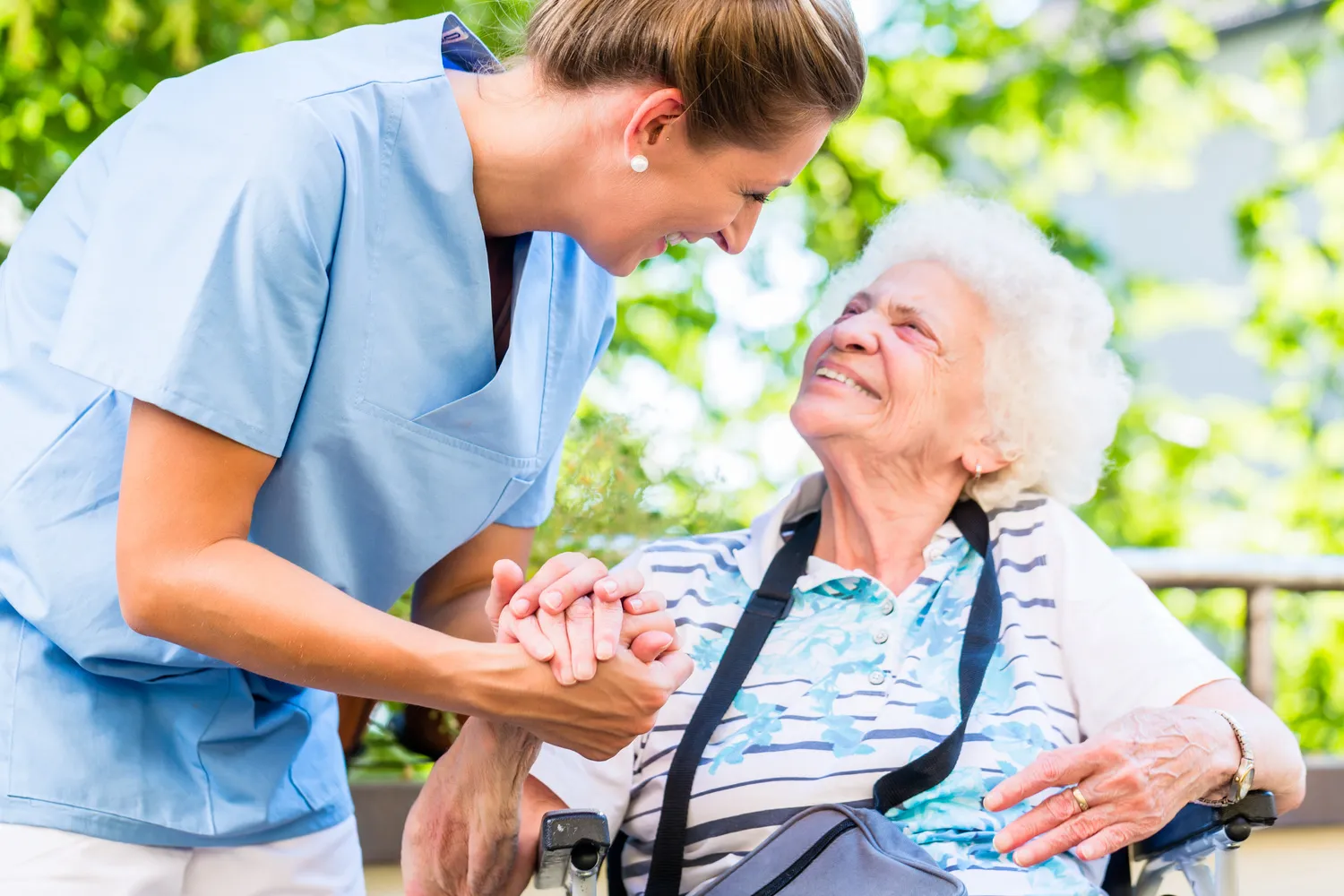 SENIOR GOLD 24 GMBH, Krankenschwester hält die Hand einer fröhlichen älteren Frau, die im Rollstuhl sitzt